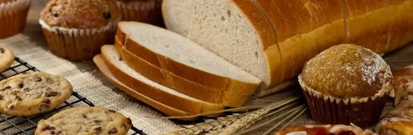 Apetyczny zestaw wypieków: bochenek jasnego chleba pokrojony w kromki, czekoladowe ciasteczka na kratce, babeczki i muffiny ustawione na rustykalnym tle z kłosami zboża. Obraz oddaje klimat domowego pieczenia i pasji do wypieków, idealny dla reklamy produktów cukierniczych i piekarniczych.