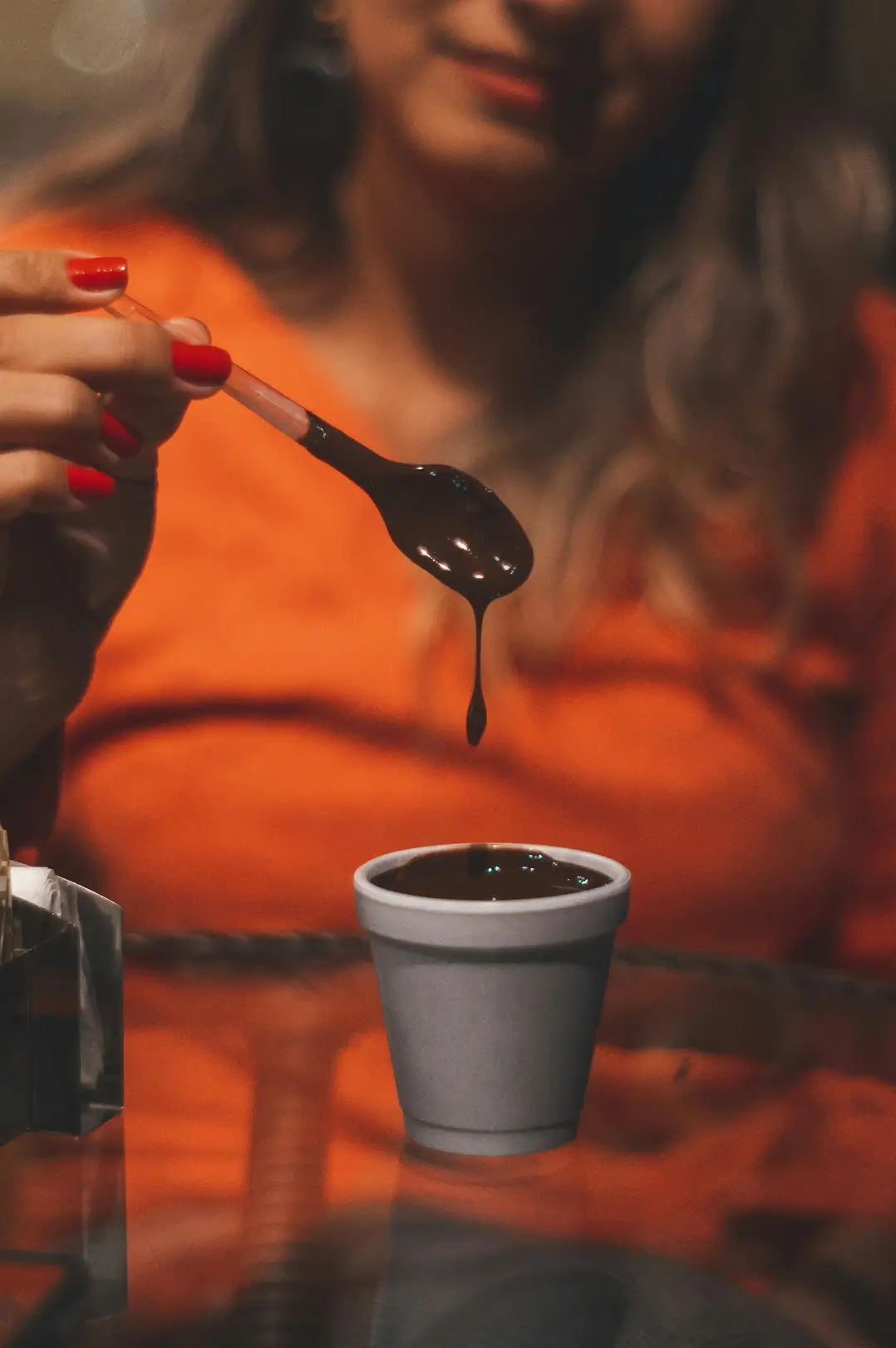 Na zdjęciu widzimy osobę trzymającą łyżeczkę nad małym kubkiem z czekoladowym fondue. Osoba ma pomalowane na czerwono paznokcie, a w tle widać jej twarz, choć jest nieco niewyraźna. Ma na sobie pomarańczowy sweter, który wyróżnia się na tle reszty obrazu. Łyżeczka jest zanurzona w czekoladzie, która kapie z jej końca. Zdjęcie jest wykonane w ciepłych tonach, co podkreśla przytulny i apetyczny charakter sceny.