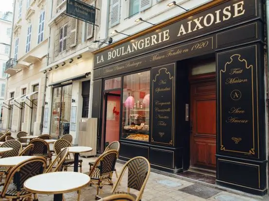Urokliwa francuska piekarnia La Boulangerie Aixoise z elegancką czarno-złotą fasadą i witryną pełną świeżych wypieków. Przed lokalem ustawione są stoliki z wiklinowymi krzesłami w stylu paryskiej kawiarni. Obraz oddaje klimat miejsca z tradycją i charakterem – idealnego dla prawdziwych smakoszy.